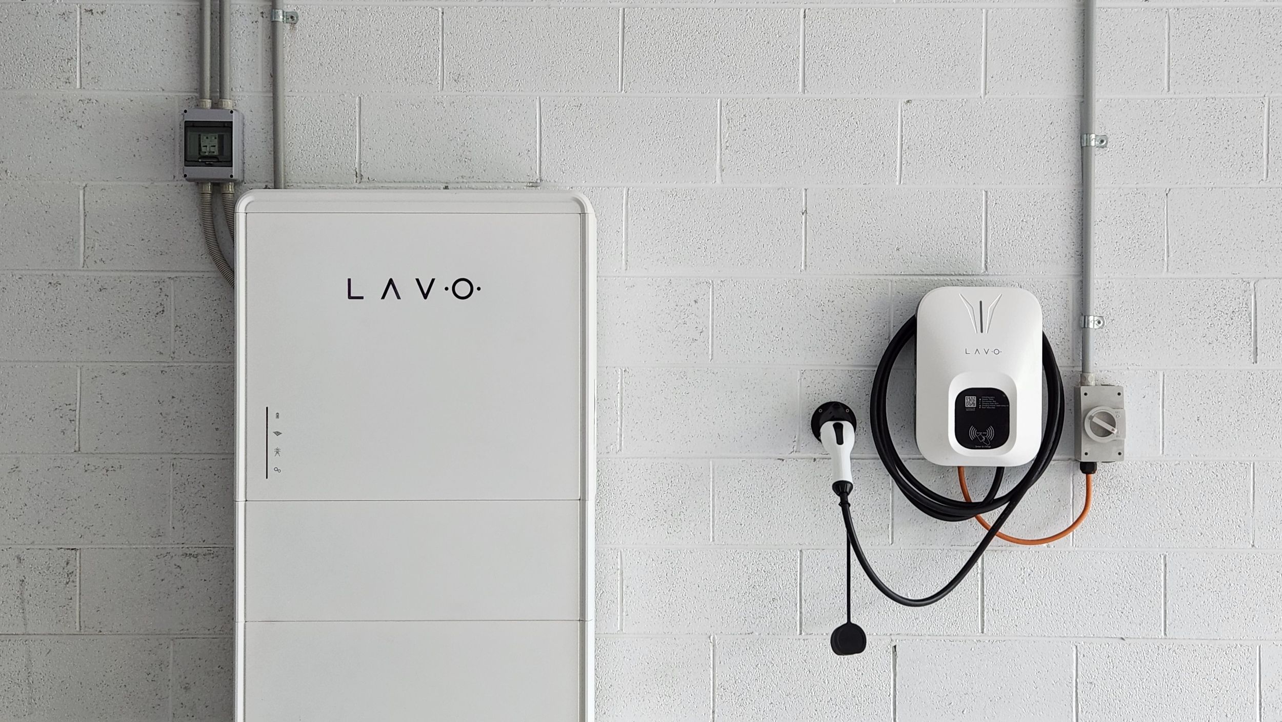 A white Lavo energy storage unit is mounted on a wall beside a charging station with a cable.