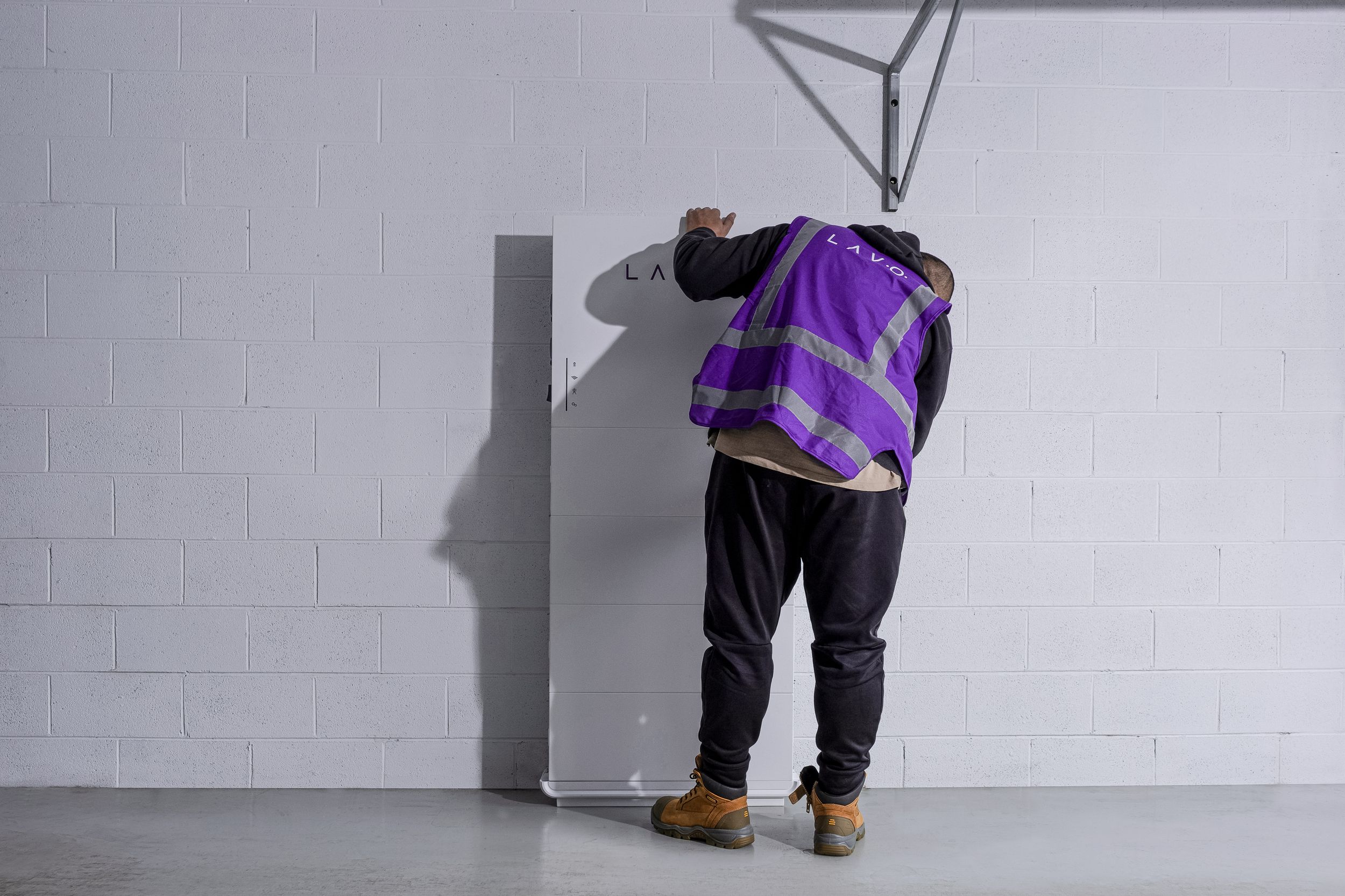 A person in a purple safety vest leans against a white wall, appearing to inspect or adjust a panel.
