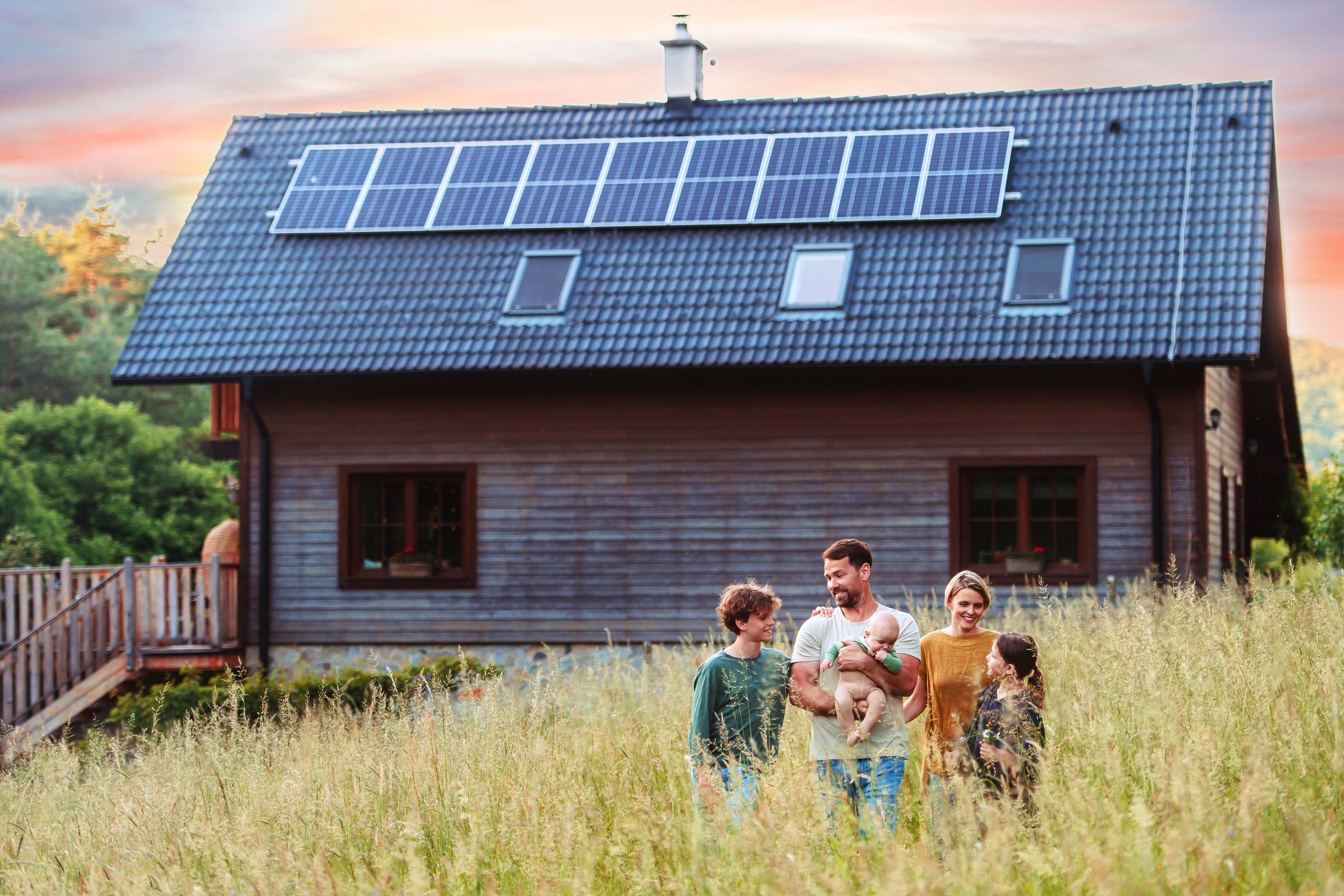A family stands in a grassy field in front of a solar-paneled house during sunset.
