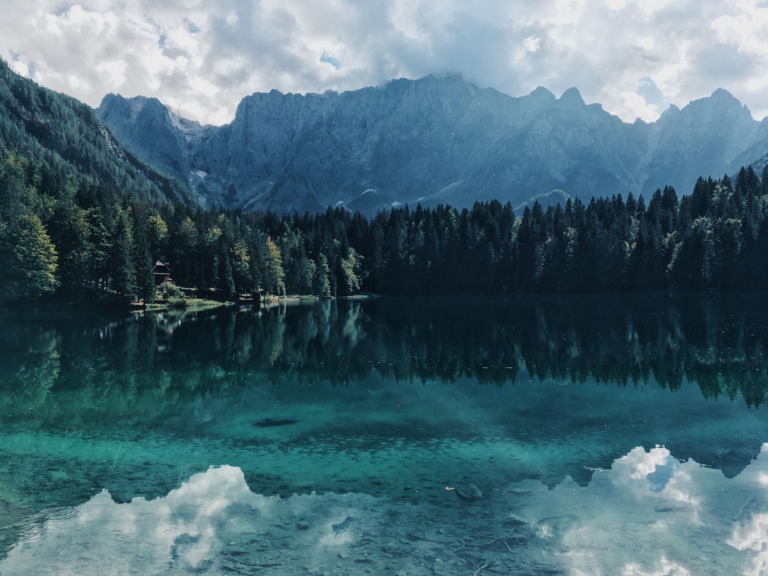 A serene lake reflects surrounding mountains and trees under a cloudy sky, creating a tranquil natural landscape.