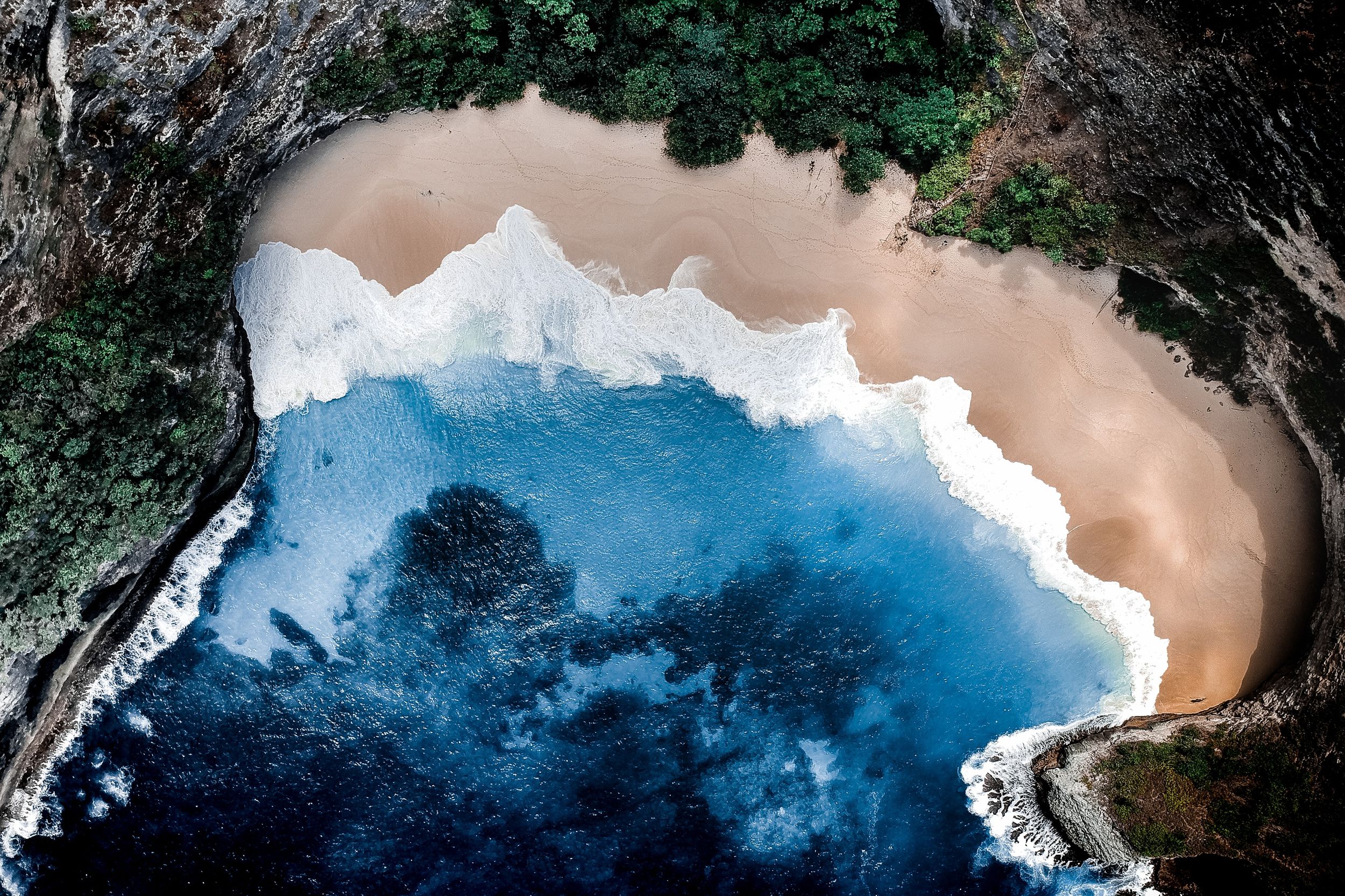 Aerial view of a secluded beach with golden sand, turquoise water, and lush greenery along the rocky coastline.