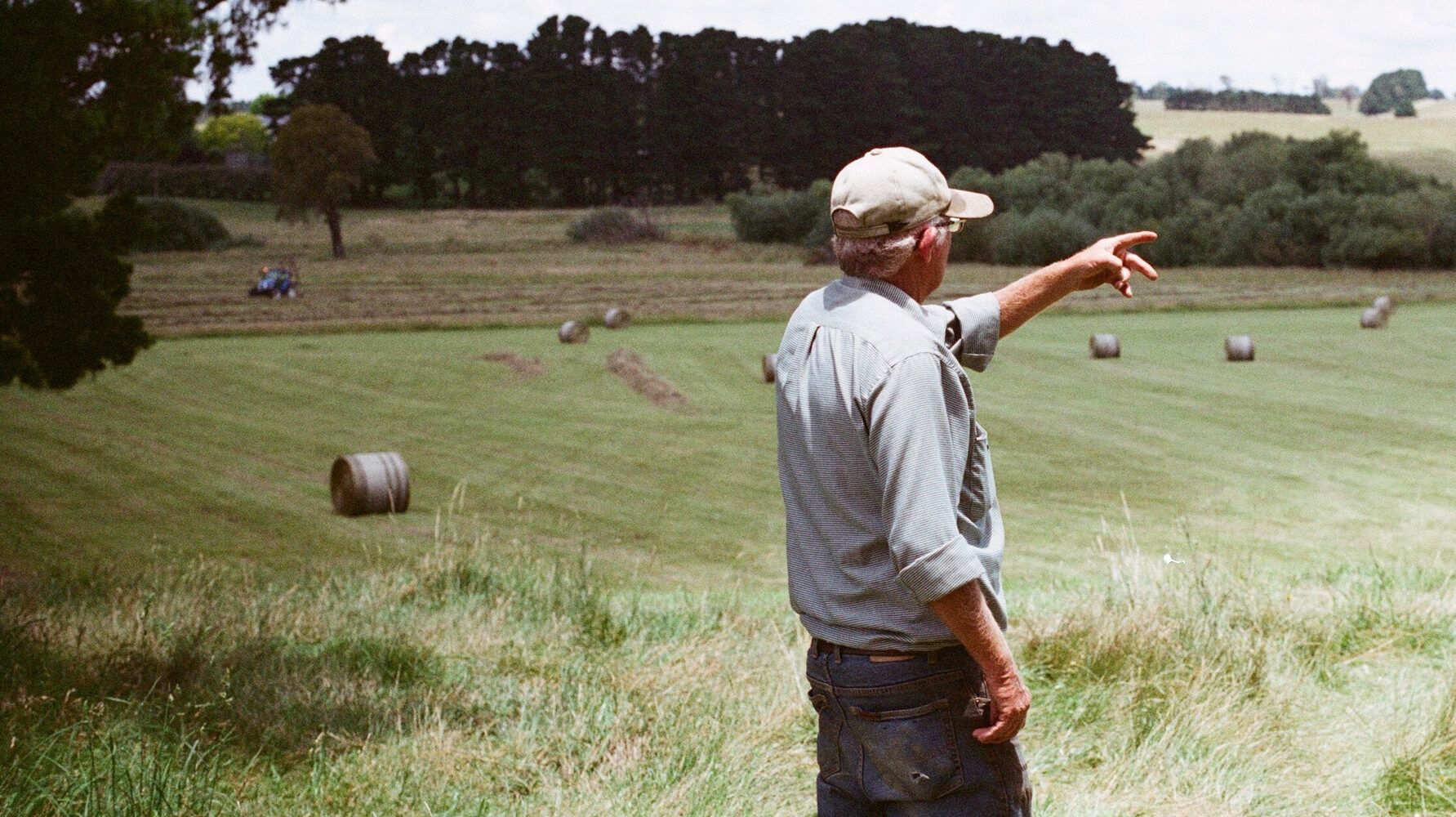 A man in a cap stands in a field, pointing towards hay bales scattered across the landscape under a cloudy sky.