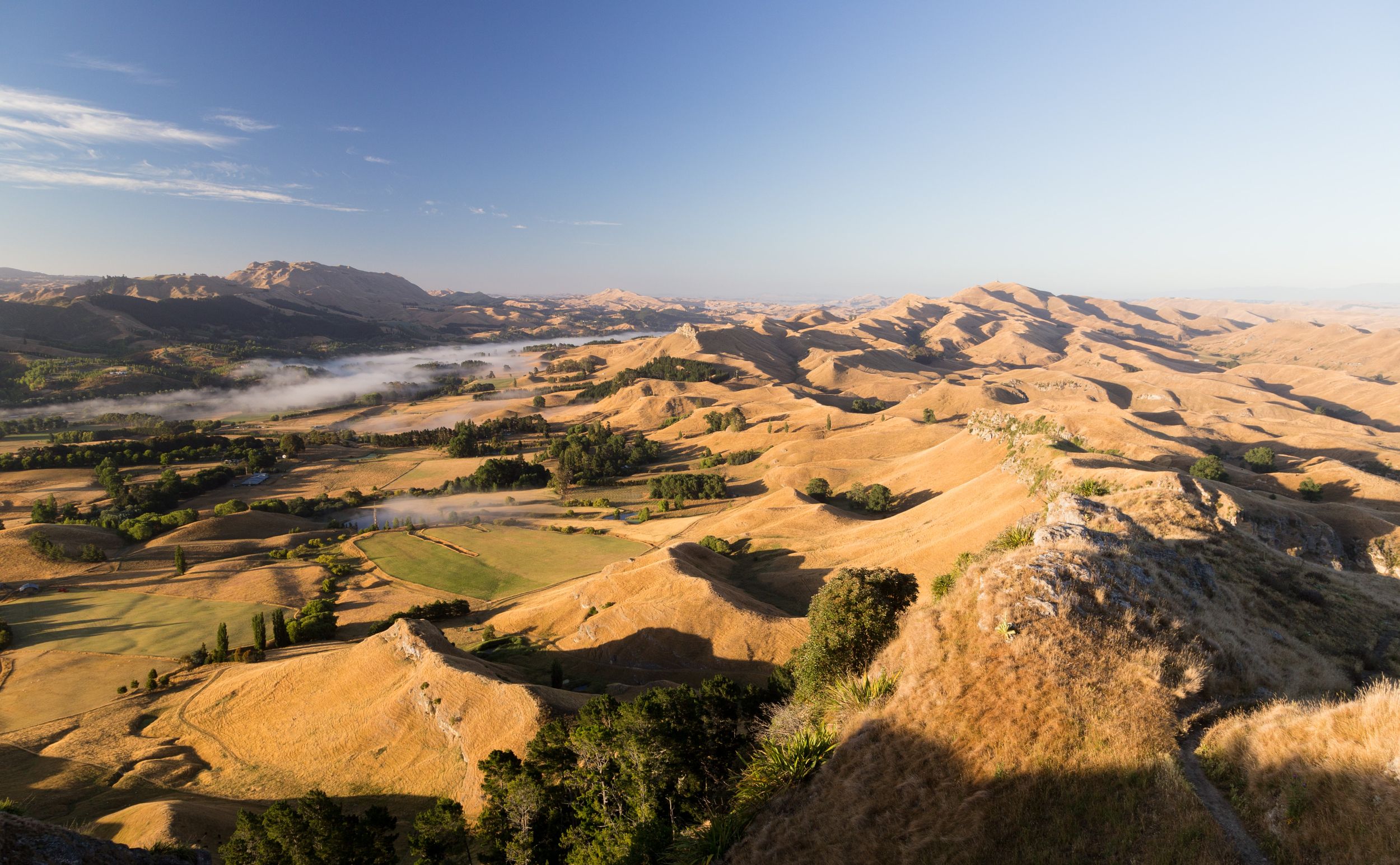A panoramic view of rolling golden hills and valleys under a clear blue sky, with mist in the distance.