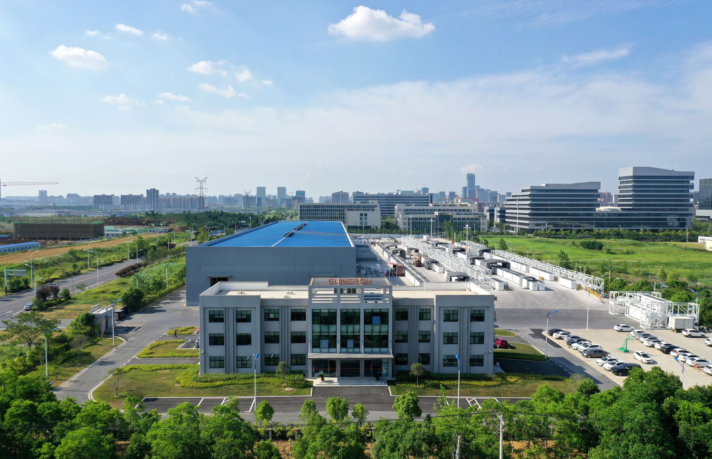 Aerial view of a modern industrial building with a blue roof, surrounded by green landscaping and other structures.